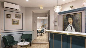 Un espace d’accueil comprenant un comptoir de réception, deux fauteuils verts, une petite table ronde et un tableau accroché au mur.