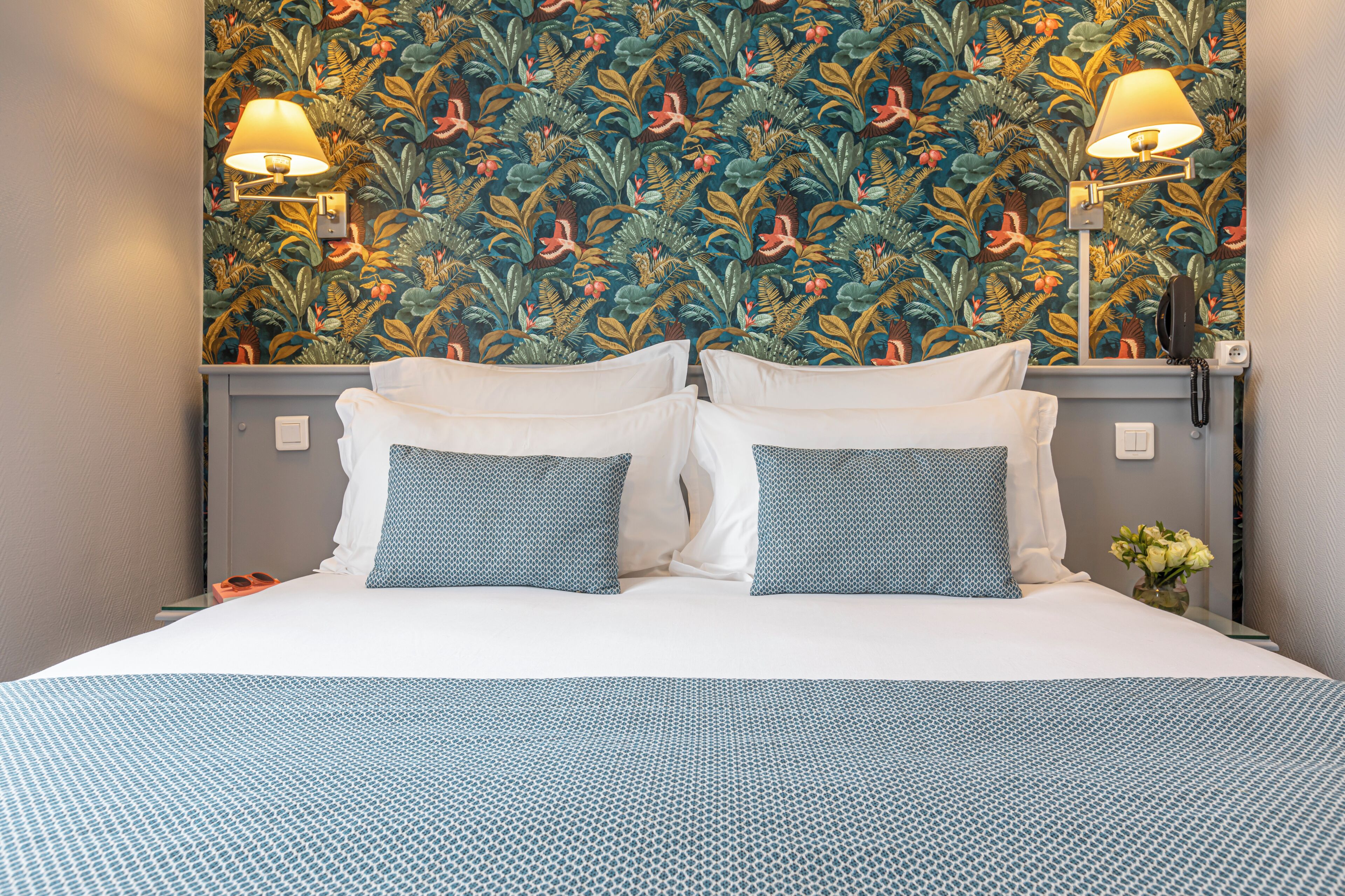 Habitación de hotel con una cama, dos lámparas de pared, un teléfono y papel tapiz con motivos florales.