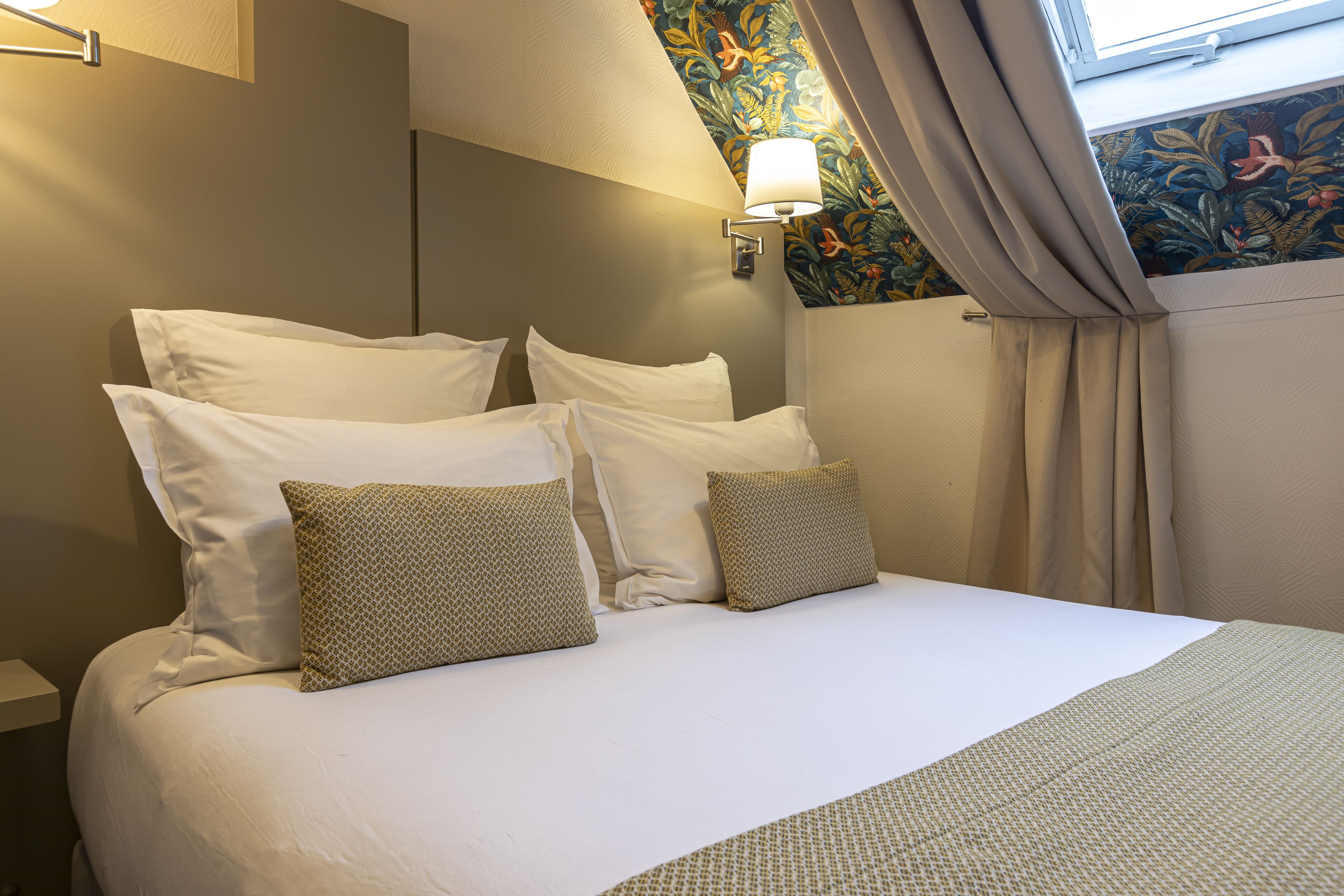 A neatly made bed with white and patterned pillows, a wall-mounted lamp, and a floral-patterned wall.