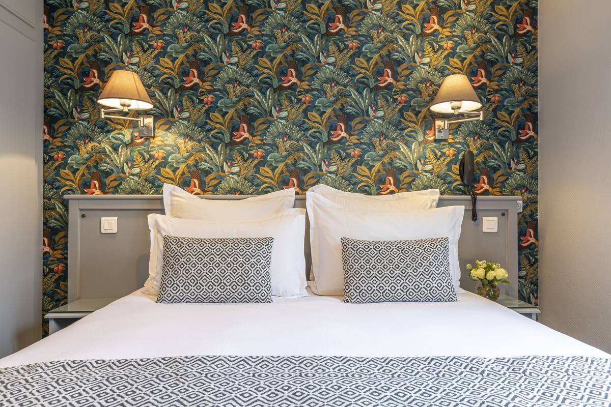 A hotel room with a bed, two patterned pillows, a nightstand with a lamp, and a floral wallpaper.
