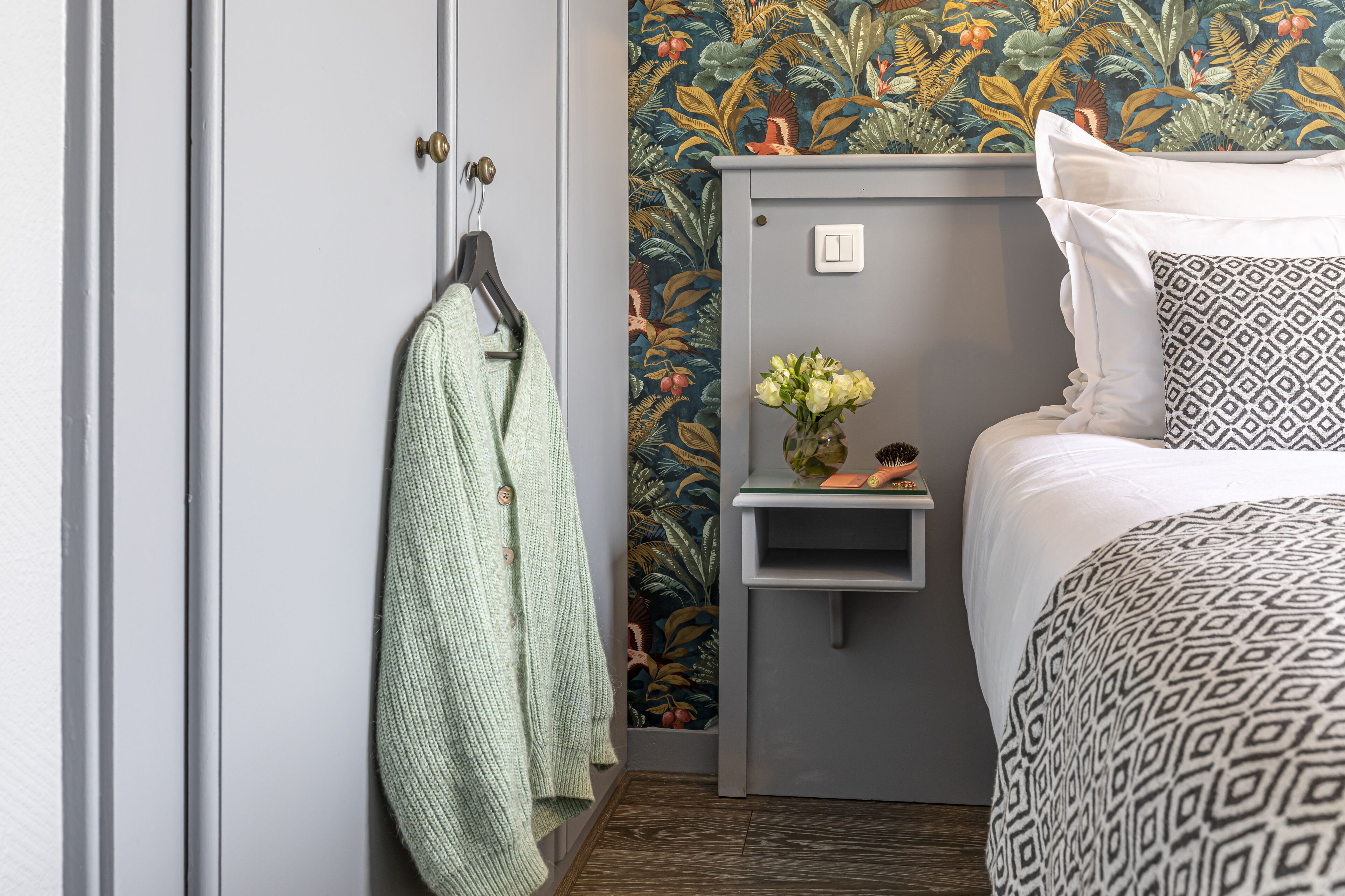 Une chambre avec une armoire grise, une serviette verte suspendue, une table de chevet ornée d’un vase de fleurs et une couvre-lit à motifs.