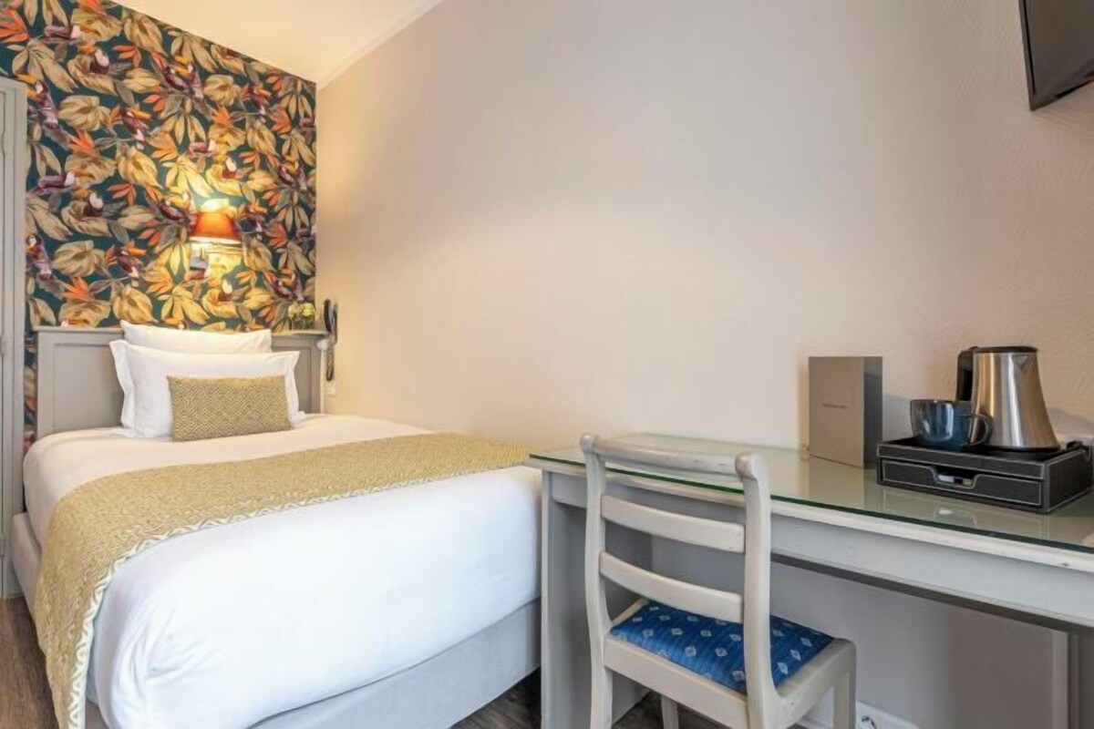 A hotel room with a bed, a desk, a chair, a coffee maker, and a wall-mounted lamp.