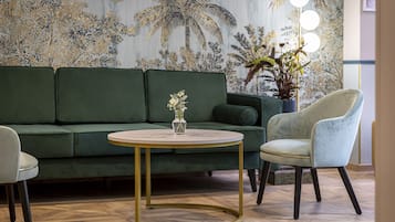 Un sofá verde, una mesa redonda con un florero y una silla azul claro en una habitación con piso de espiga y un mural tropical.