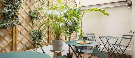 Un patio con una mesa redonda y sillas, rodeado de plantas en macetas y un muro de celosía de madera.
