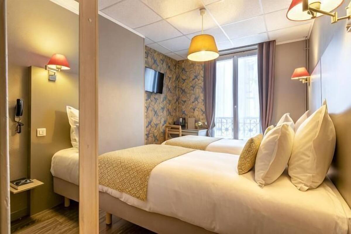 A hotel room with a bed, bedside table, lamp, TV, and a window with curtains.