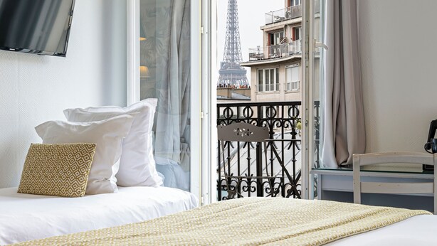 Une chambre d’hôtel avec un lit, une télévision et une vue sur la Tour Eiffel, visible par la porte ouverte du balcon.