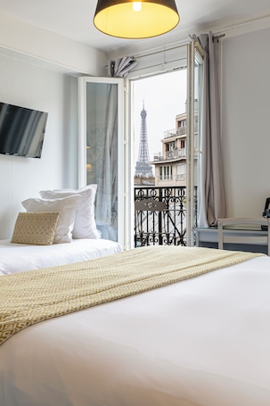 A hotel room with a bed, a TV mounted on the wall, and a view of the Eiffel Tower through an open balcony door.