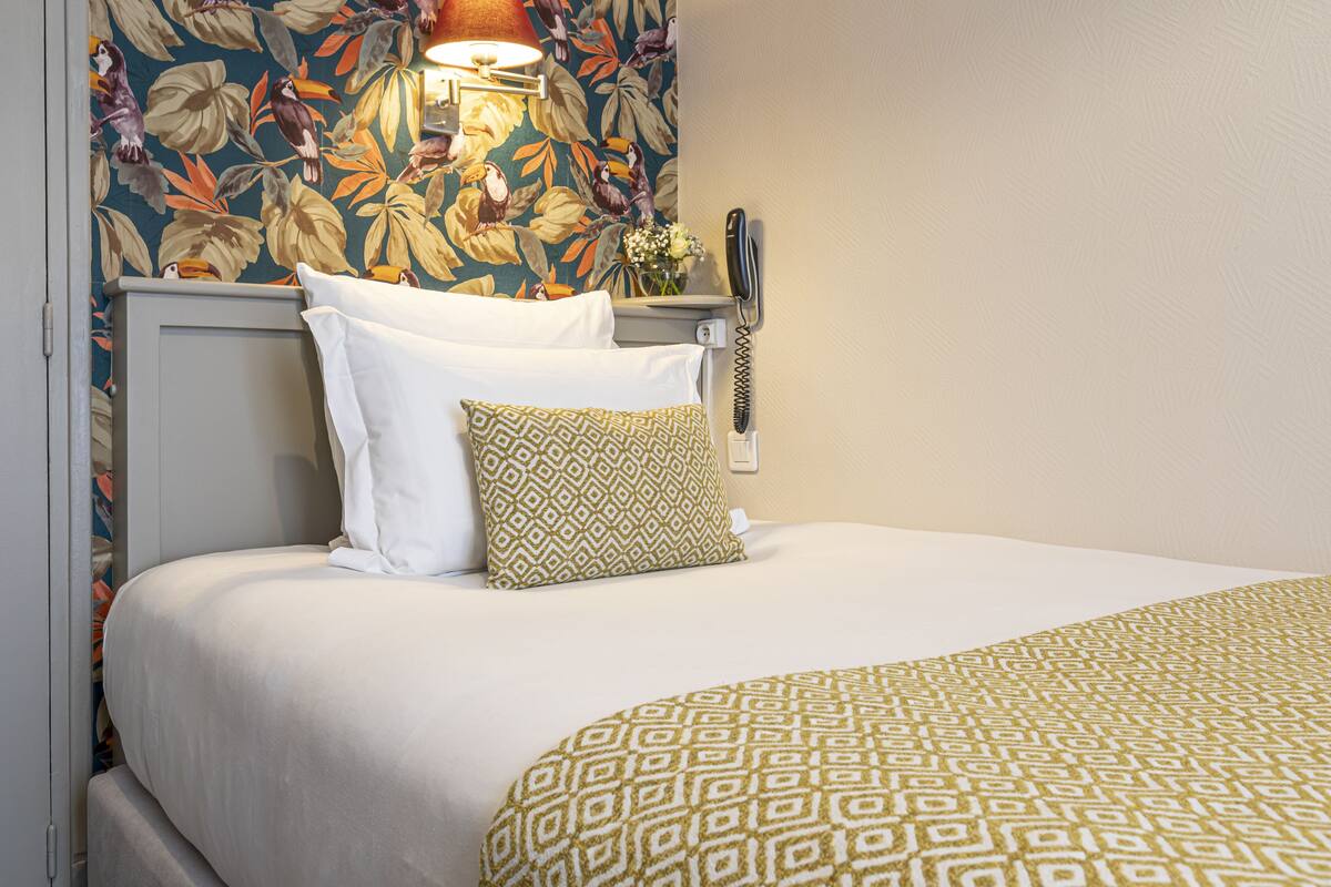 A neatly made bed with white and patterned pillows, a floral wallpaper, and a bedside telephone.