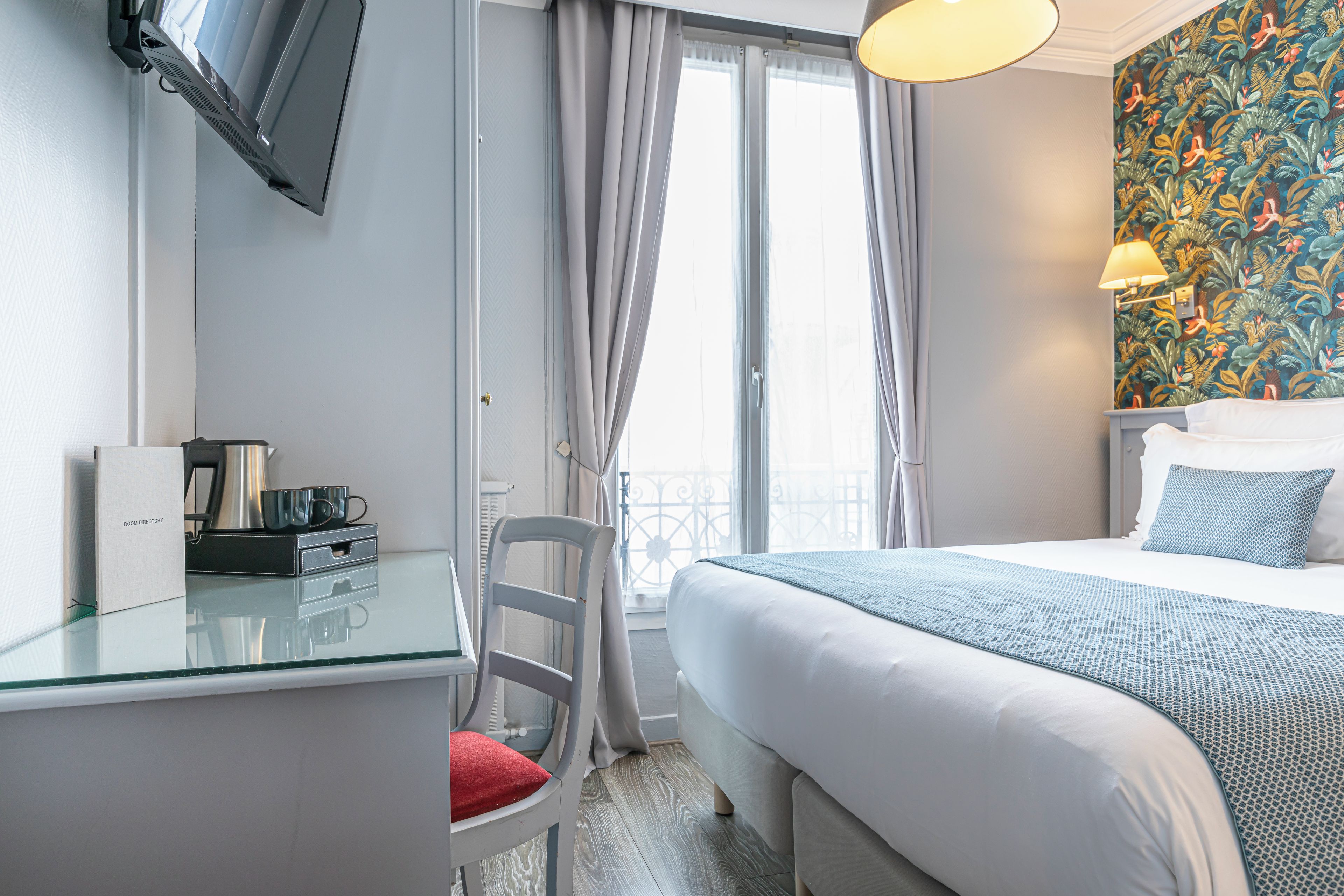A hotel room with a bed, a desk with a chair, a TV mounted on the wall, and a window with curtains.