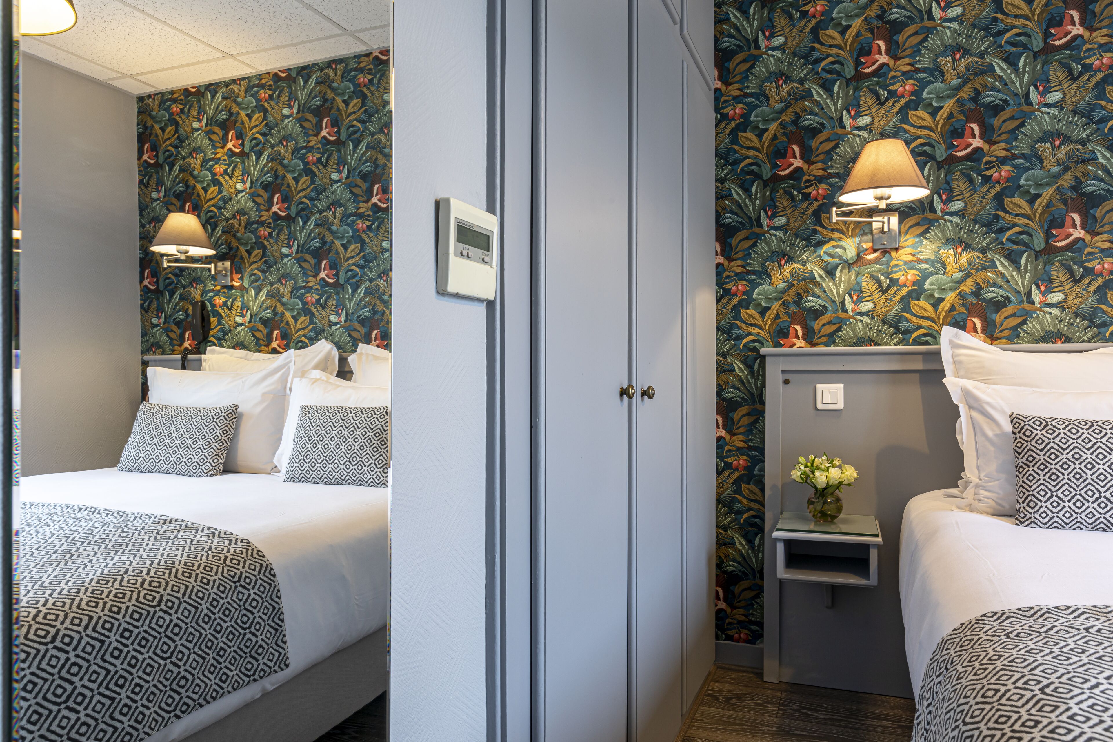 A hotel room with two beds, a wardrobe, a nightstand with a lamp, and a floral wallpaper.