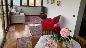 A living room with a sofa, red chair, and a table with a floral tablecloth. There are three paintings on the wall and a large window with a view of the sea. - MaLu House - (Messina)