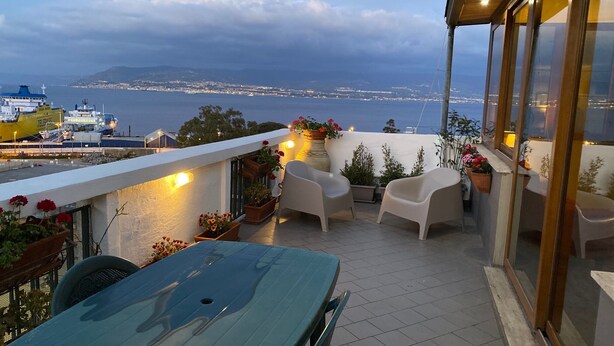 A terrace with a table and chairs, potted plants, and a view of the sea and distant cityscape. - MaLu House - (Messina)