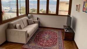 A room with a sofa, a rug, a TV, and a view of a body of water and mountains through large windows. - MaLu House - (Messina)