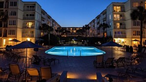 Una piscina amb gandules i ombrelles, envoltada d'edificis de diversos pisos amb balcons i il·luminació exterior.