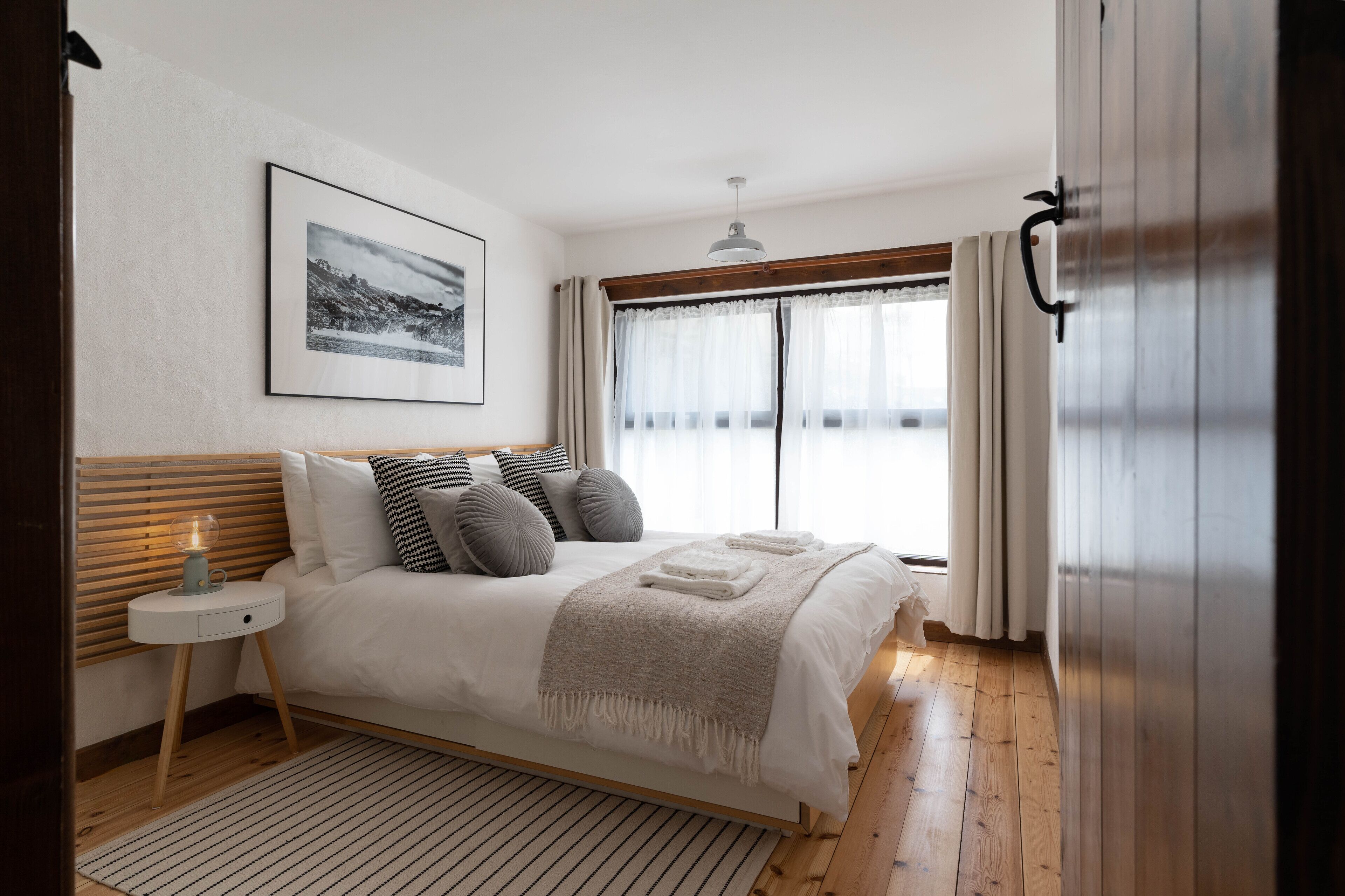 Un dormitorio con una cama de madera, una mesita de noche con lámpara, un cuadro enmarcado en la pared y una ventana con cortinas.