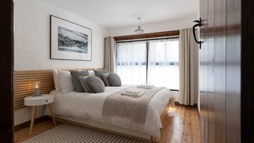 Un dormitorio con una cama de madera, una mesita de noche con lámpara, un cuadro enmarcado en la pared y una ventana con cortinas.