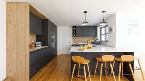 A modern kitchen with dark cabinetry, a wooden accent wall, a central island with bar stools, and pendant lighting. - Delightful Seaside Hideaway 5 Mins to Beach (Chichester)