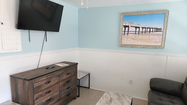 A room with a TV mounted on the wall, a wooden dresser, a marble table, a cushioned sofa, and a framed picture of a pier. - Harrison Hotel Unit F
Pet Friendly 
Ground Level (Murrells Inlet)