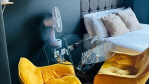 A modern bedroom with a bed, two yellow chairs, a glass table, a lamp, and a painting on the wall. - Holland Rd Studios Close to Olympia London (London)