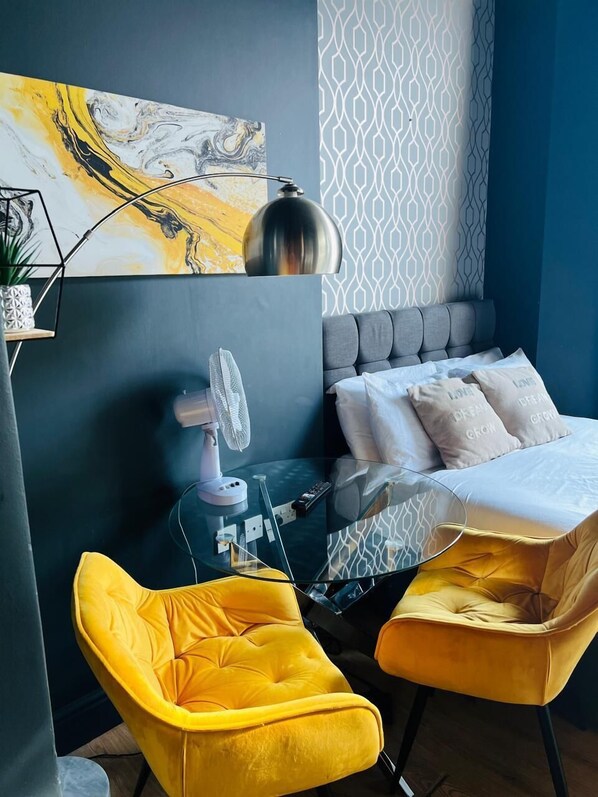 A modern bedroom with a bed, two yellow chairs, a glass table, a lamp, and a painting on the wall. - Holland Rd Studios Close to Olympia London (London)