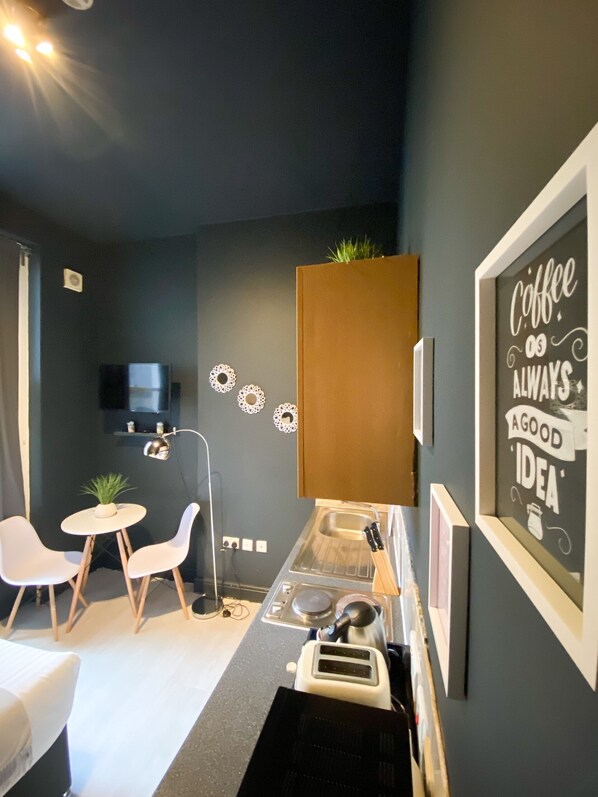 A compact kitchen with a coffee machine, two white chairs, a small table with a plant, and a wall-mounted TV.