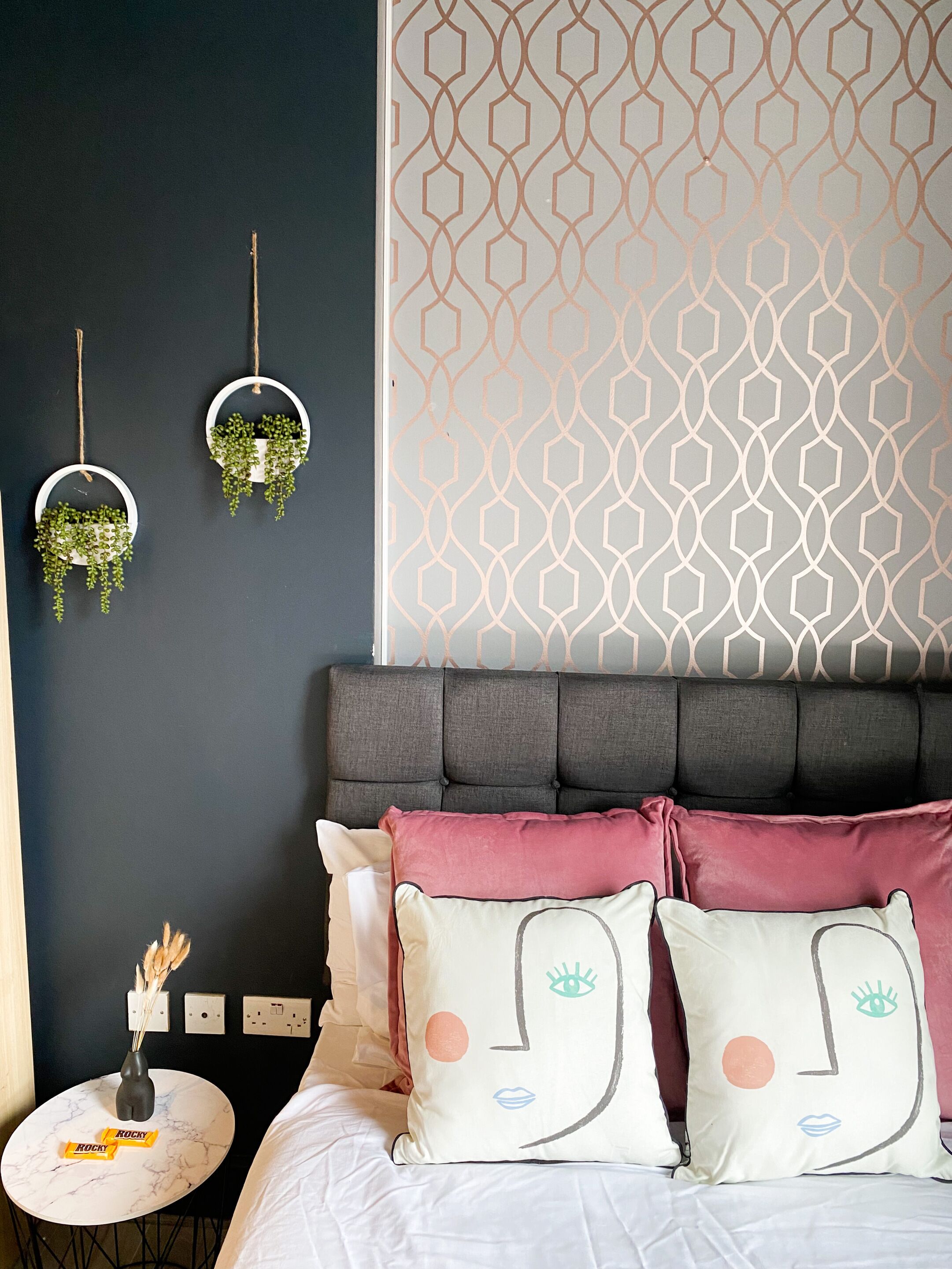 Un dormitorio con pared oscura, papel tapiz de diseño geométrico, cabecera capitonada y dos macetas colgantes circulares con follaje.