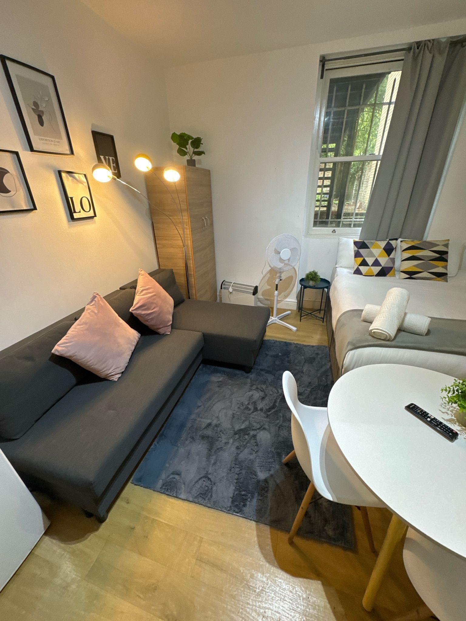 Ein modernes Wohnzimmer mit einem Ecksofa, einem kleinen Esstisch und einem weißen Stuhl. Eine Stehlampe, ein Standventilator und ein Fenster mit Vorhängen sind ebenfalls vorhanden.