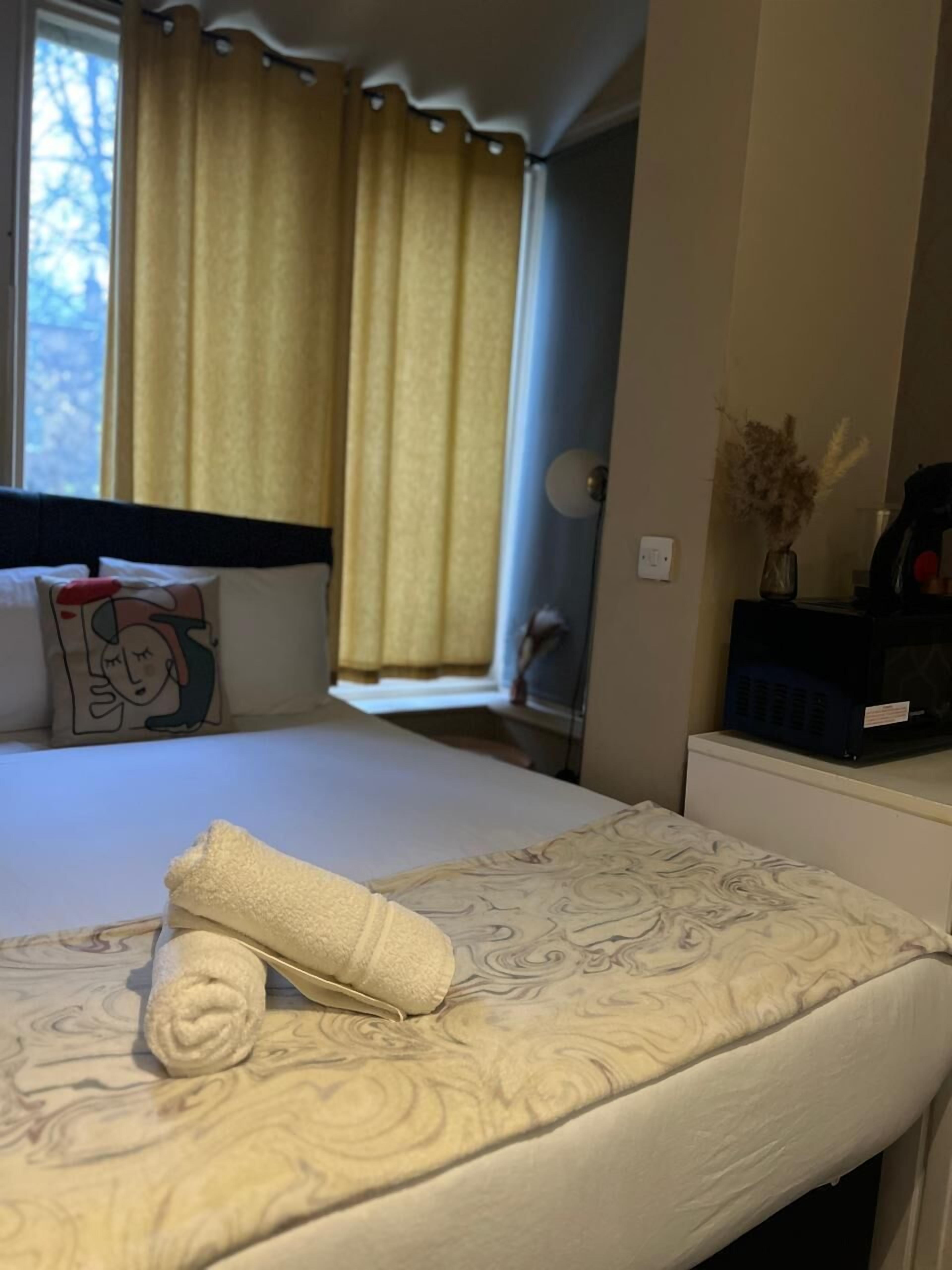 Une chambre d’hôtel avec un lit, une fenêtre avec des rideaux, une table de chevet ornée d’un vase de fleurs séchées et un radiateur fixé au mur.