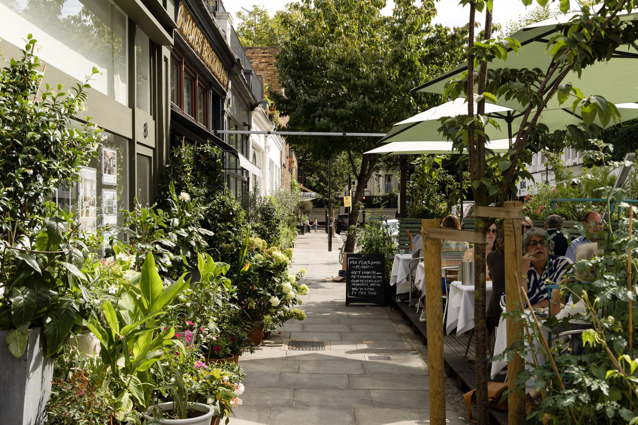Een smalle, open corridor met plantenbakken en tafels, doet denken aan een café of restaurant.