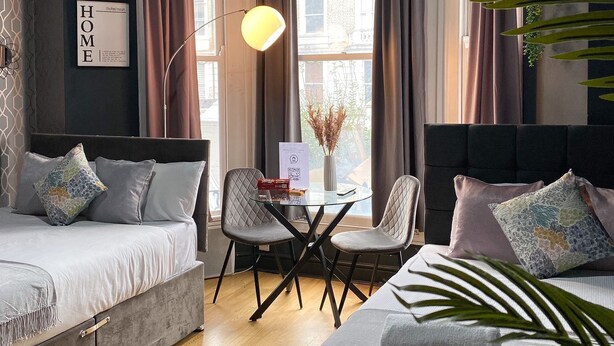 A modern hotel room with two beds, a small table with a lamp, and a view of buildings through large windows. - Holland Rd Studios Close to Olympia London (London)