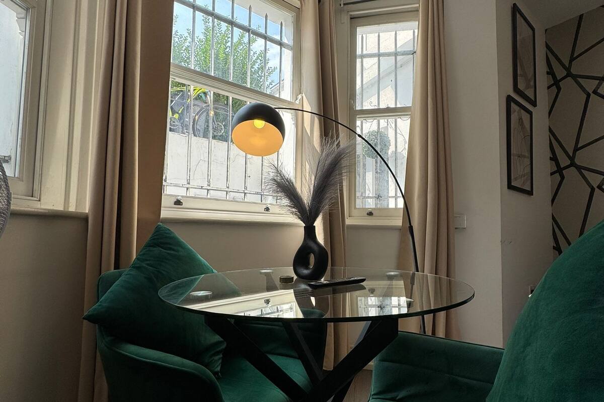 A room with a glass table, two green chairs, a lamp, and a window with curtains.
