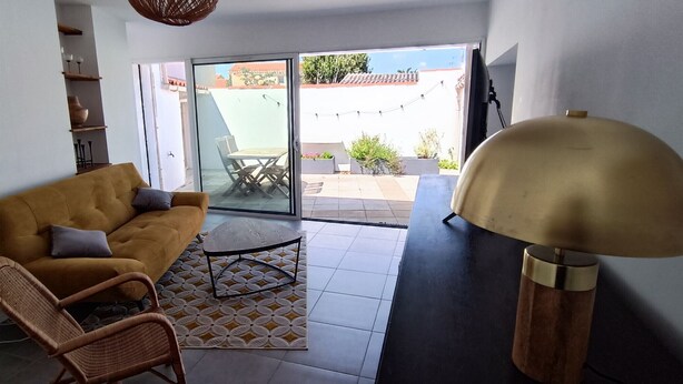Sala de estar con sofá, una silla, una mesa y una lámpara. Hay un ventanal corredizo que da a un área exterior con una mesa y sillas.