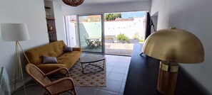 Sala de estar con sofá, una silla, una mesa y una lámpara. Hay un ventanal corredizo que da a un área exterior con una mesa y sillas.
