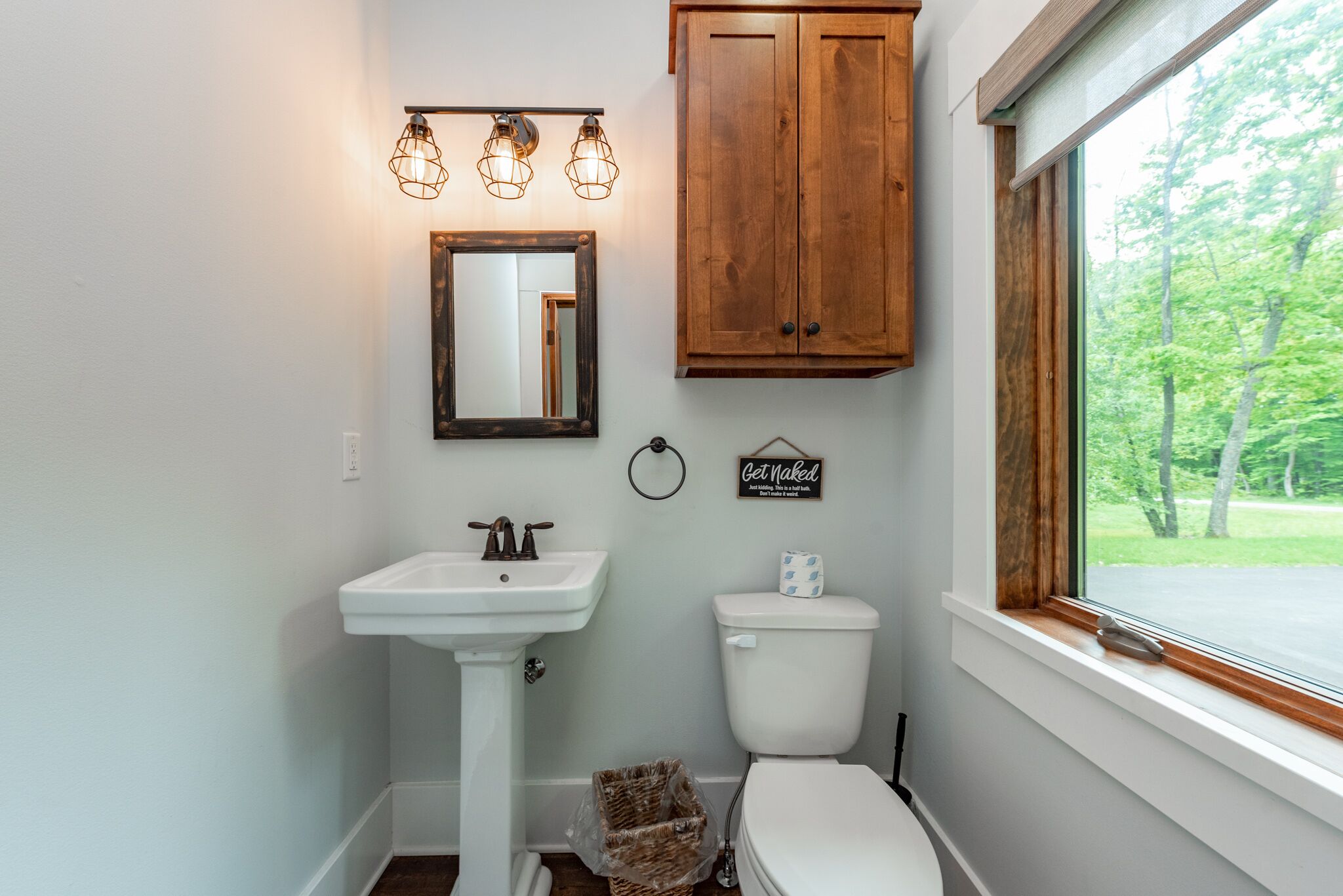 A bathroom with a white sink, wooden cabinet, and a toilet. There is a window with a view of trees.