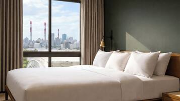 Habitación de hotel con una cama grande, una mesita de noche, una lámpara y vistas a un paisaje urbano a través de un ventanal amplio.