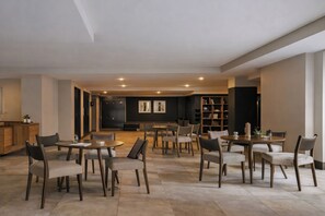 A spacious dining area with multiple round tables and chairs, a bookshelf, and a dark-toned wall with framed artwork.