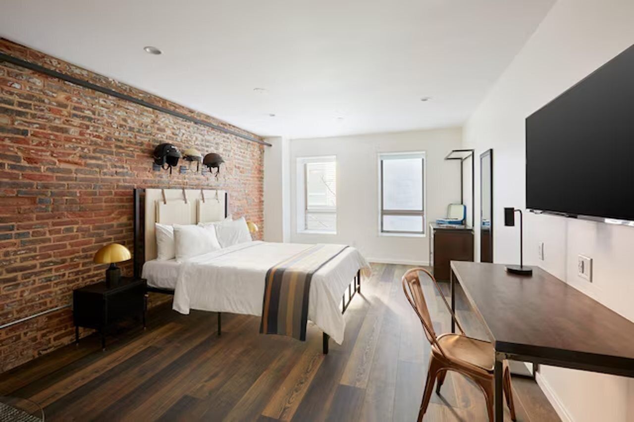 Habitación de hotel moderna con pared de ladrillo, una cama grande, piso de madera, un escritorio y un televisor de pantalla plana.