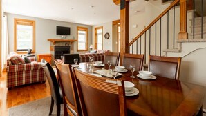 Comedor con una mesa de madera dispuesta para cuatro personas, rodeada de sillas. Una escalera con barandillas de madera conduce a un piso superior.