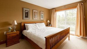 Un dormitorio con cama de madera, dos mesitas de noche, una mesita auxiliar, una ventana con cortinas y tres cuadros enmarcados en la pared.