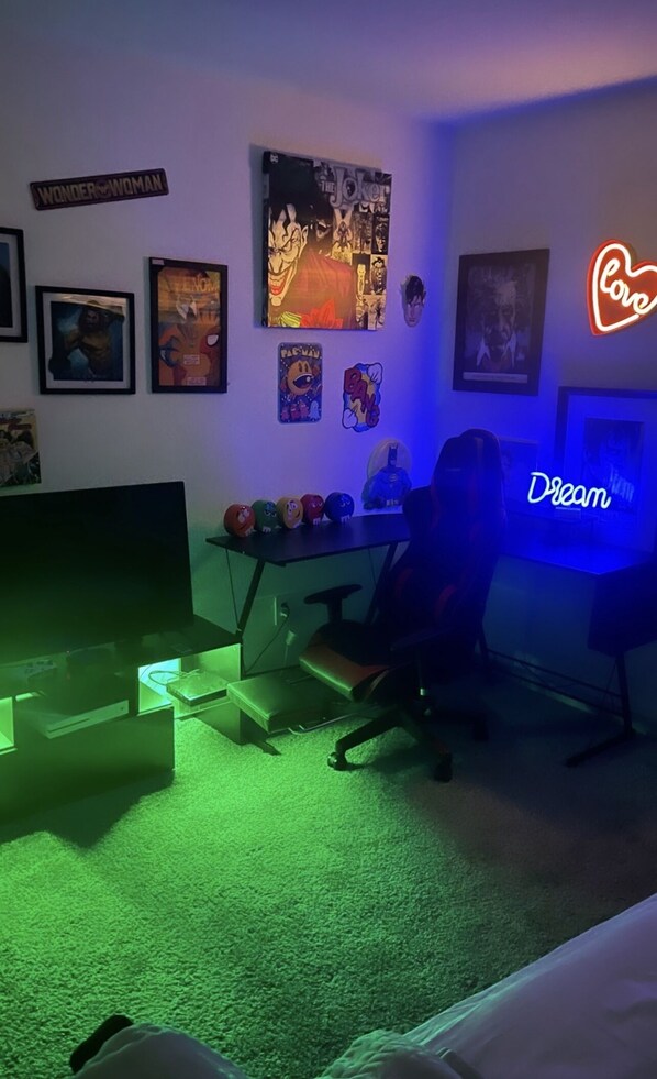 Relax in a cozy space with a gaming desk, computer, and chair, surrounded by fun posters and a vibrant neon sign.
