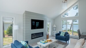 A modern living room with a fireplace, a flat-screen TV, and a view of the lake. - Make memories at this unique family friendly beach-front home in Mashpee  (Mashpee)