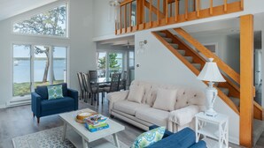 A spacious living area with a large window overlooking a lake, a staircase with wooden railings, and a dining area with a glass table and chairs. - Make memories at this unique family friendly beach-front home in Mashpee  (Mashpee)