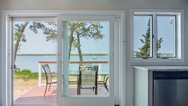 A modern kitchen with a dishwasher, a sliding glass door leading to a deck with outdoor seating, and a view of a lake with boats. - Make memories at this unique family friendly beach-front home in Mashpee  (Mashpee)