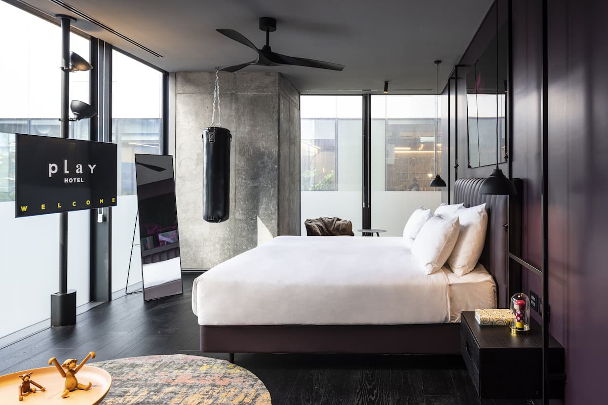 A modern hotel room with a large bed, a ceiling fan, a punching bag, and a TV displaying "PLAY HOTEL WELCOME".