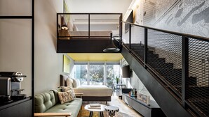 Een modern loft appartement met een hoge plafond, een slaapkamer op een entresol, een trap met een metalen leuning, en een groot raam met uitzicht op de omgeving.