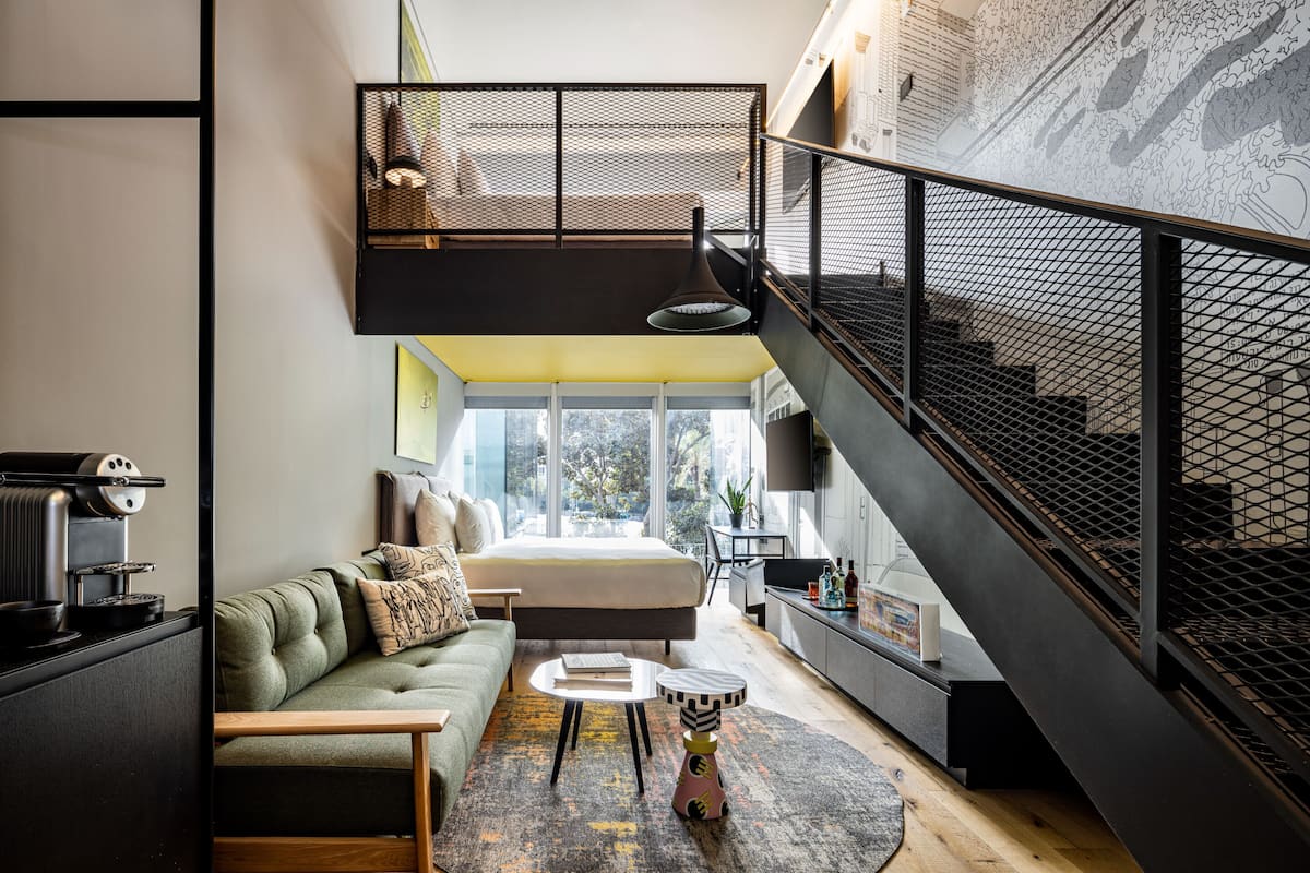 A modern loft apartment with a high ceiling, a mezzanine bedroom, a staircase with a metal railing, and a large window providing a view of the outdoors.