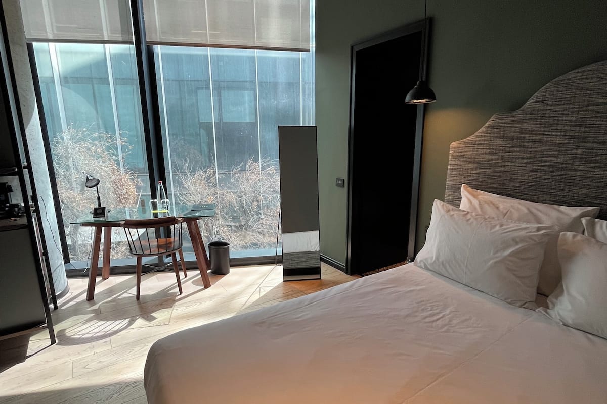 A hotel room with a large bed, a desk with a chair, a standing lamp, and a view of trees through large windows.