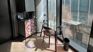 Een moderne kamer met een glazen tafel, een stoel, een kast met een geometrisch patroon en een groot raam met uitzicht op een gebouw en bomen.