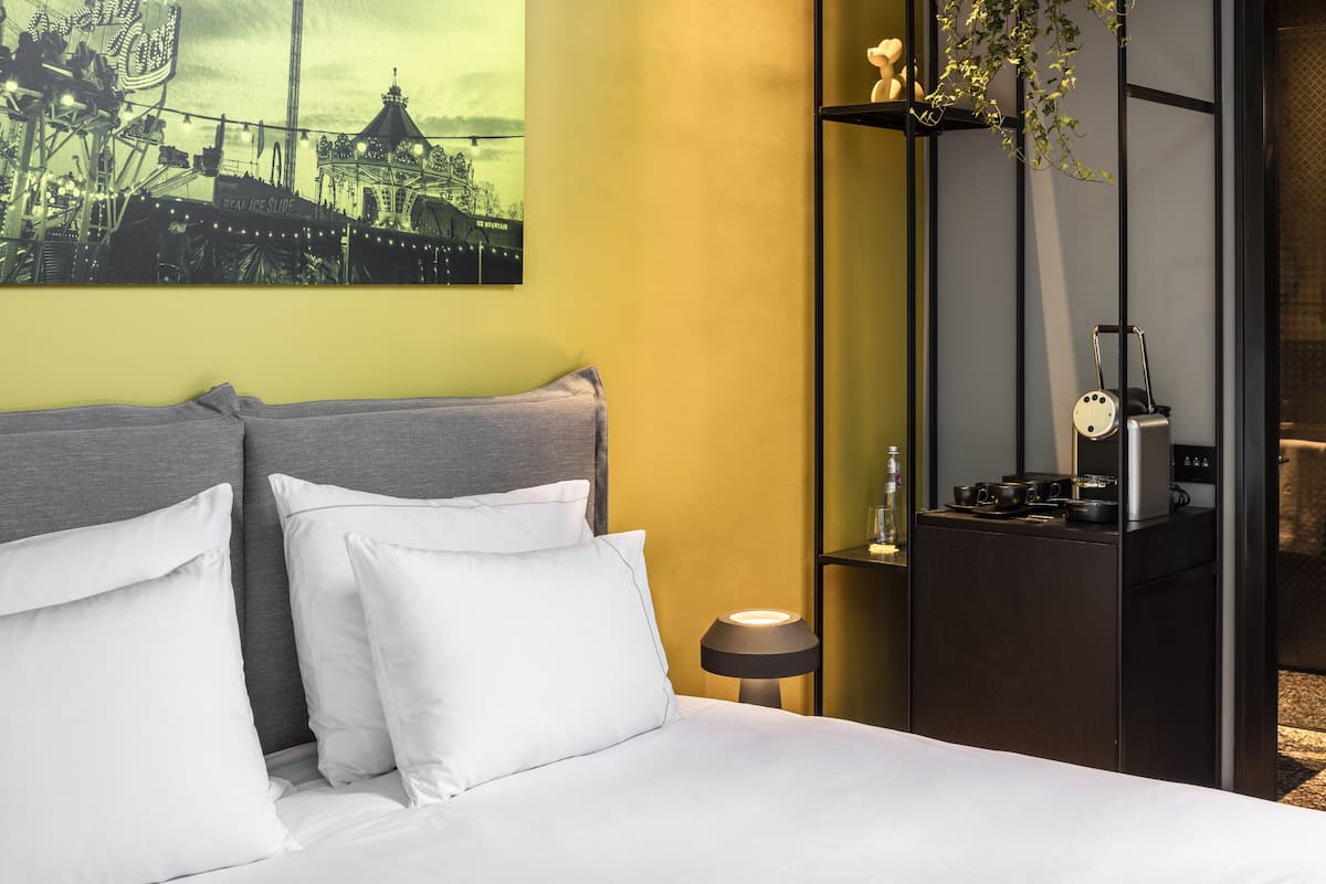 A hotel room with a bed, pillows, a nightstand with a lamp, a wall-mounted shelf with a plant, and a poster of a cityscape.