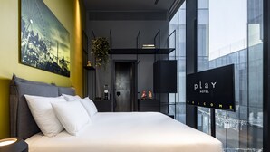 Een moderne hotelkamer met een groot bed, een stadslandschap schilderij, een digitaal welkomstbord en een uitzicht door grote ramen.
